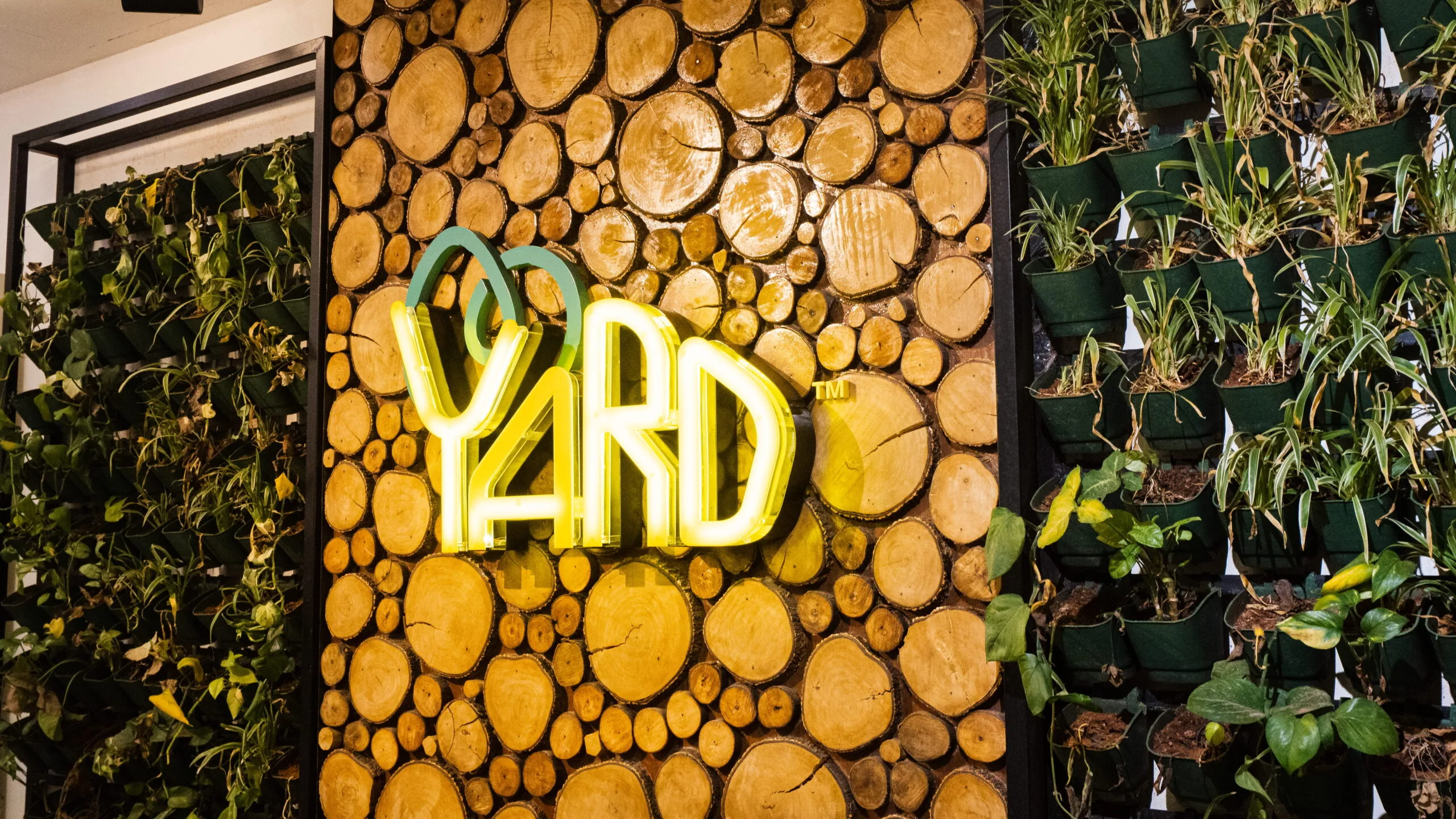 The Yard coworking space entrance featuring a wood log accent wall, indoor vertical garden, and illuminated logo, showcasing a modern eco-friendly workspace design.