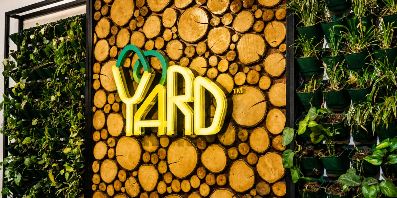 The Yard coworking space entrance featuring a wood log accent wall, indoor vertical garden, and illuminated logo, showcasing a modern eco-friendly workspace design.