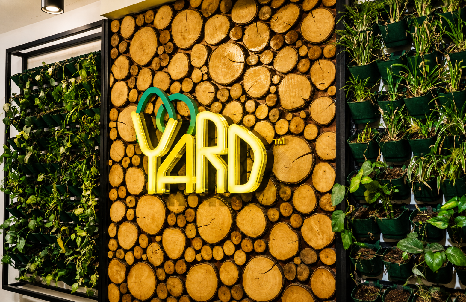 The Yard coworking space entrance featuring a wood log accent wall, indoor vertical garden, and illuminated logo, showcasing a modern eco-friendly workspace design.