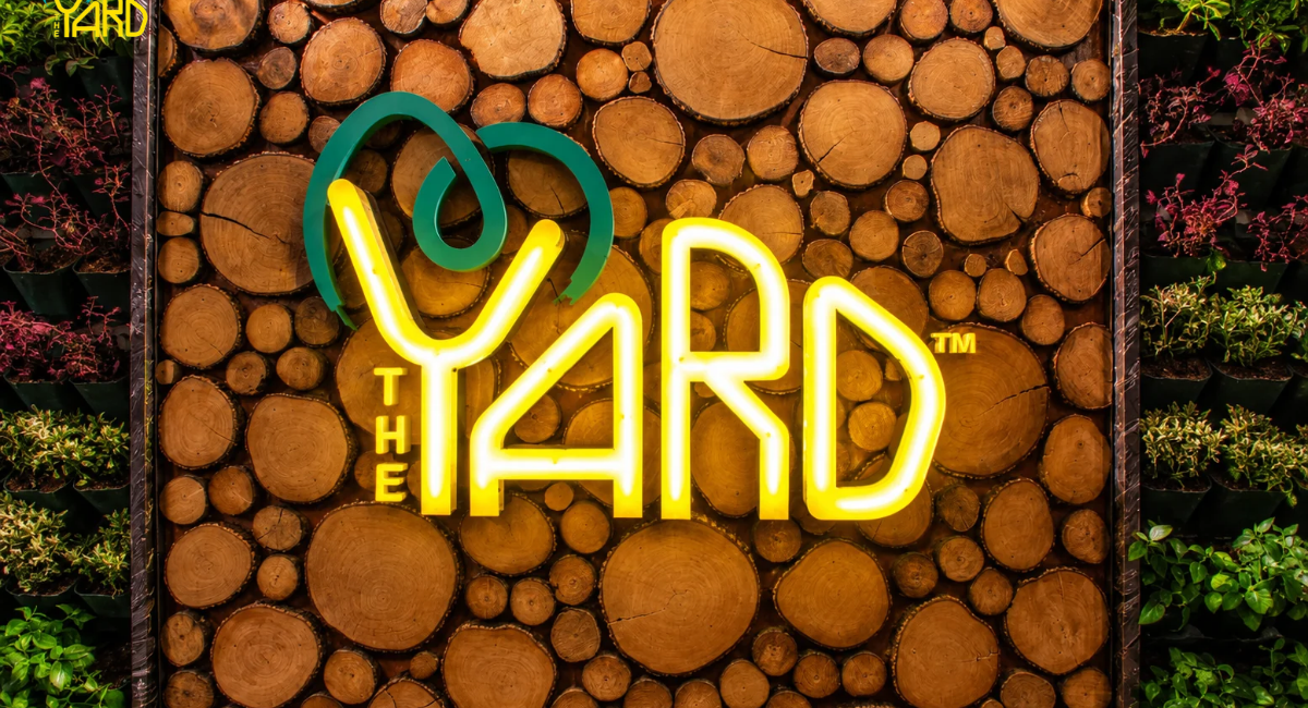 The Yard illuminated logo on a wooden wall surrounded by plants.