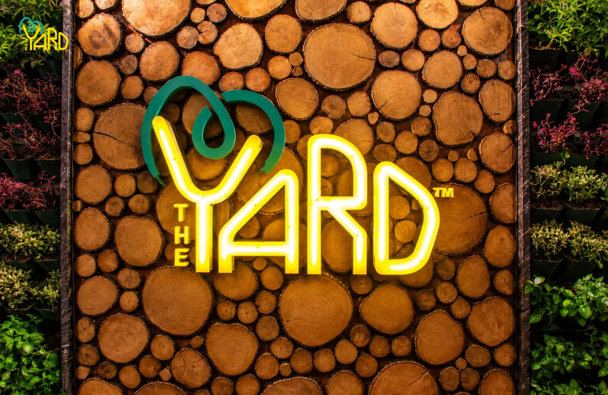The Yard illuminated logo on a wooden wall surrounded by plants.